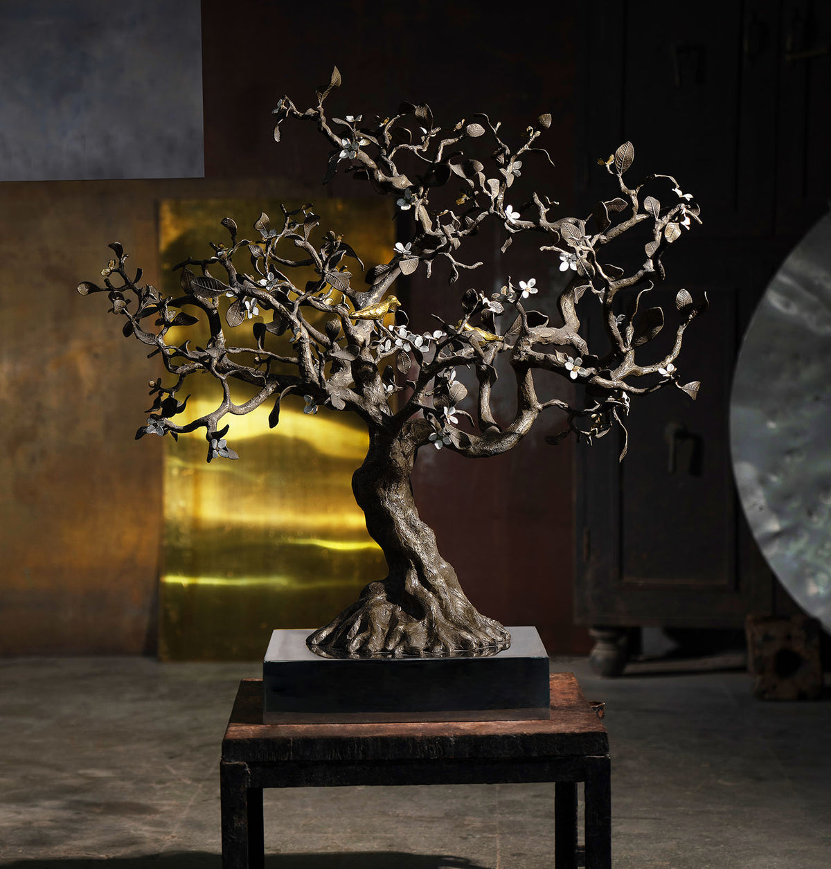 A sculptural metal bonsai-style tree sculpture with textured branches and small white blossoms, displayed on a pedestal table in a dim interior setting.