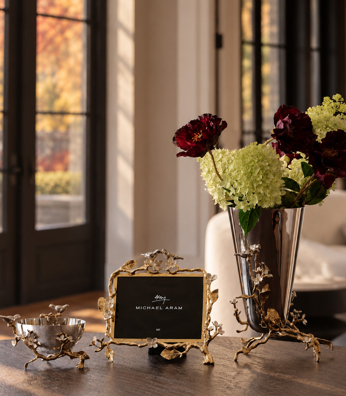 An ornate gold leaf table decor set displayed on a wooden table in a sunlit room. The set includes a gold leaf picture frame holding a black sign, a matching gold leaf bowl, and a gold leaf vase holding burgundy and light green hydrangeas.