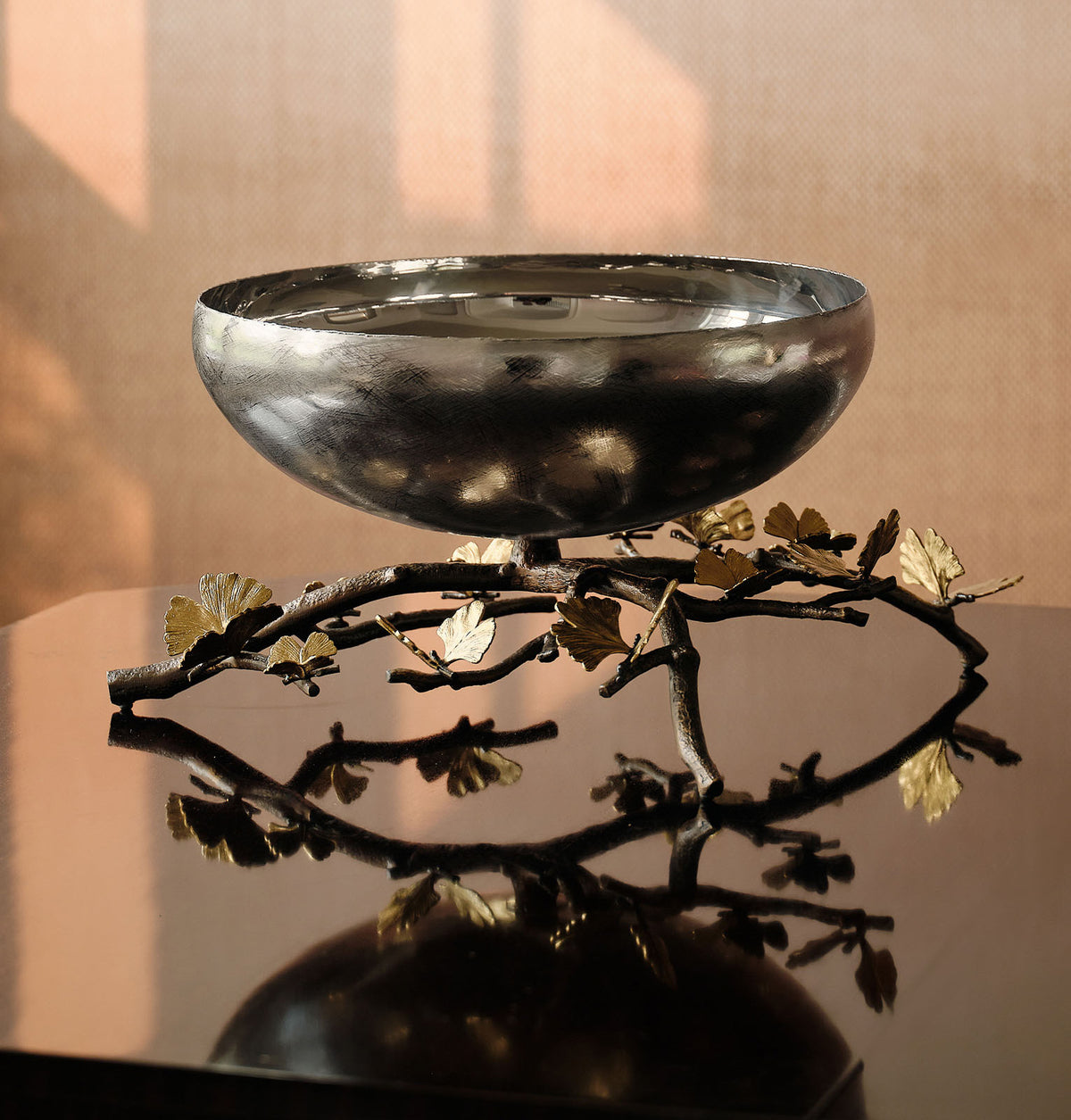 A polished silver bowl resting on a decorative branch-shaped stand with gold-toned leaf accents, photographed indoors against a warm beige wall with soft window shadows.