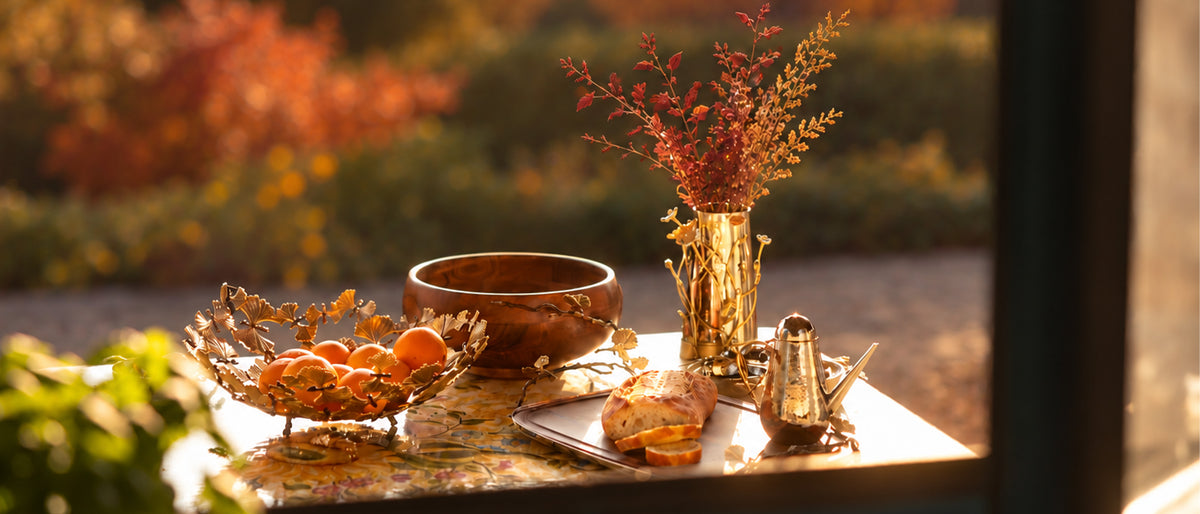 A styled autumnal tabletop scene with a gold bowl of oranges, a wooden bowl, a gold vase with dried branches, and a small metal pitcher, set against a warm blurred outdoor background.