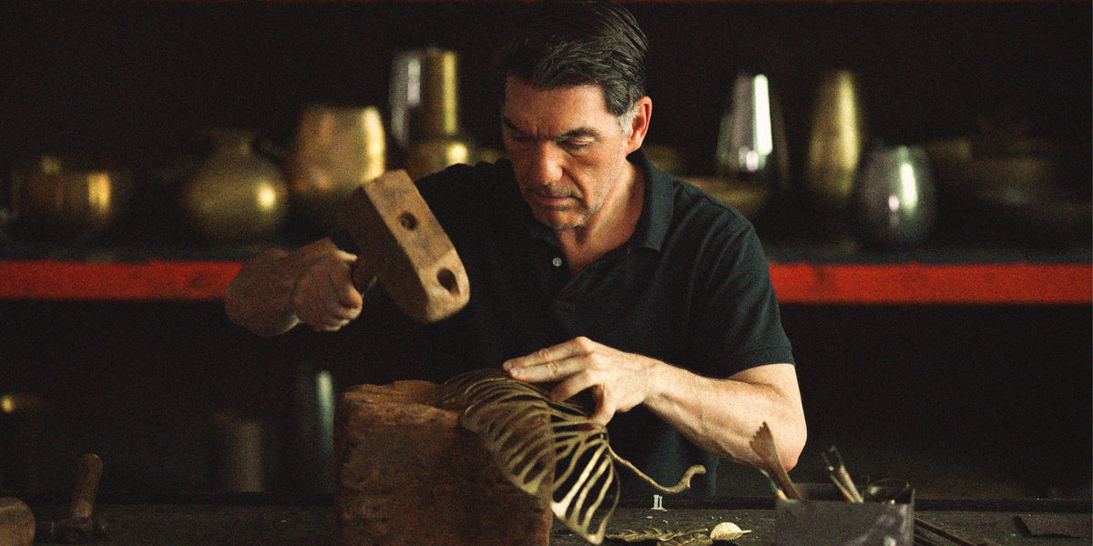 Michael Aram is shown in a dark workshop setting, using a wooden mallet to strike a carved wooden object on a workbench. Shelves with metallic vessels are blurred in the background.