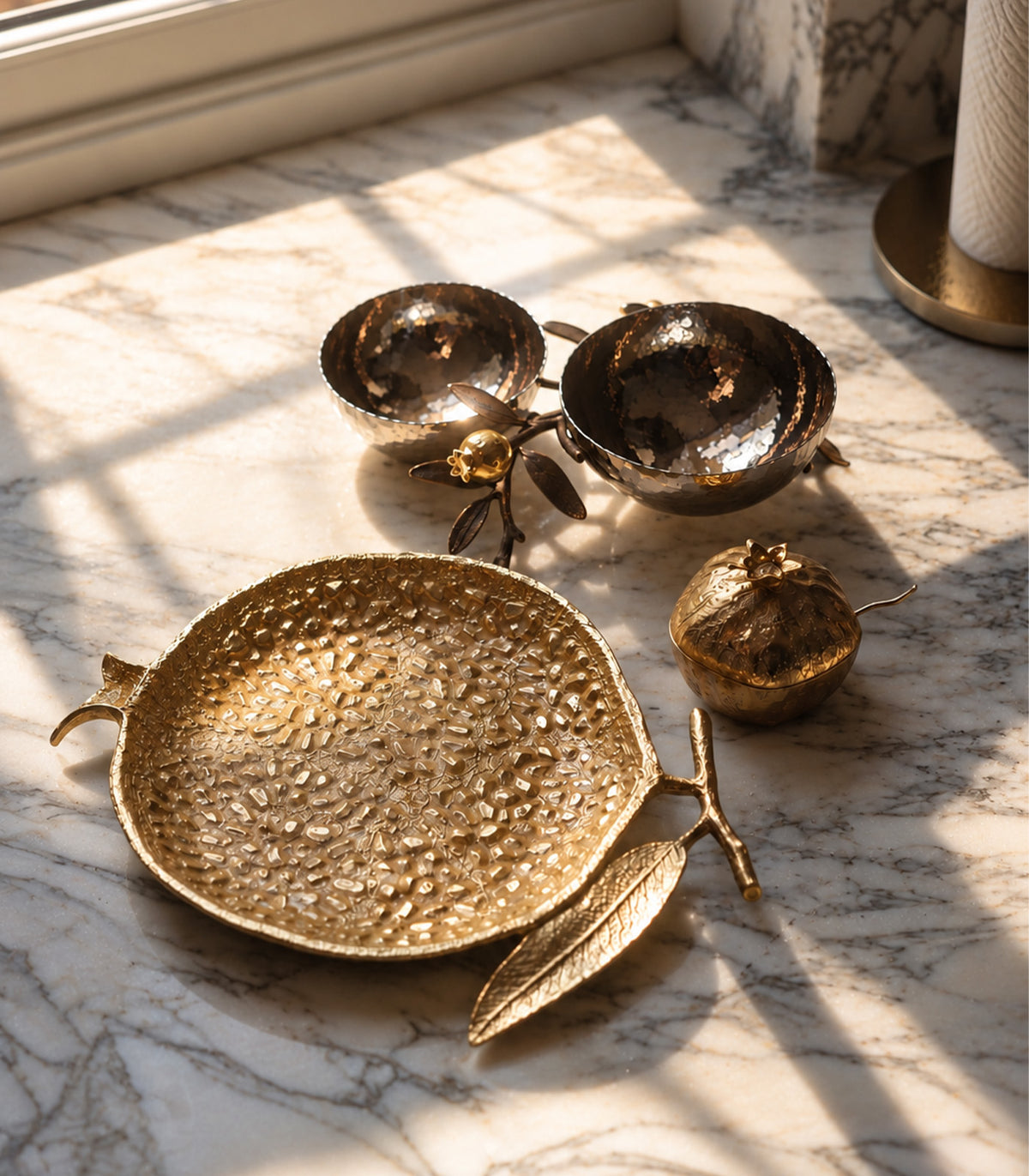 A set of decorative metallic home accessories including a hammered gold tray, two small gold bowls, and a gold pomegranate-shaped object, arranged on a marble surface with warm sunlight and window shadows.